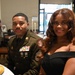 Two individuals are seated at a formal dining table, posing for a photo. The man on the left is wearing a U.S. Army dress uniform with multiple service ribbons, rank insignia consistent with a Senior Non-Commissioned Officer (Master Sergeant or Sergeant First Class), and a U.S. Army shoulder patch. The woman on the right is wearing an elegant off-shoulder black dress, suggesting this is a formal military event or banquet.