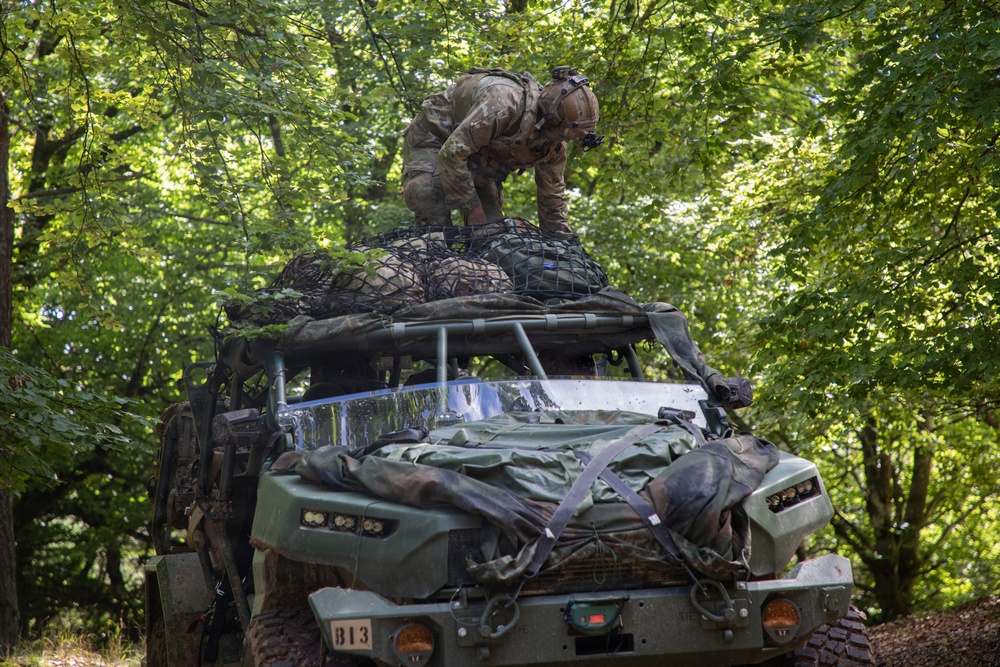 A soldier in full combat gear and helmet is crouched on top of a military light tactical vehicle, appearing to secure or adjust cargo held down by a cargo net. The vehicle, numbered '813', is parked in a wooded area with dense green foliage overhead. Additional equipment and camouflage materials are strapped to the front of the vehicle.