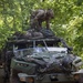 A soldier in full combat gear and helmet is crouched on top of a military light tactical vehicle, appearing to secure or adjust cargo held down by a cargo net. The vehicle, numbered '813', is parked in a wooded area with dense green foliage overhead. Additional equipment and camouflage materials are strapped to the front of the vehicle.