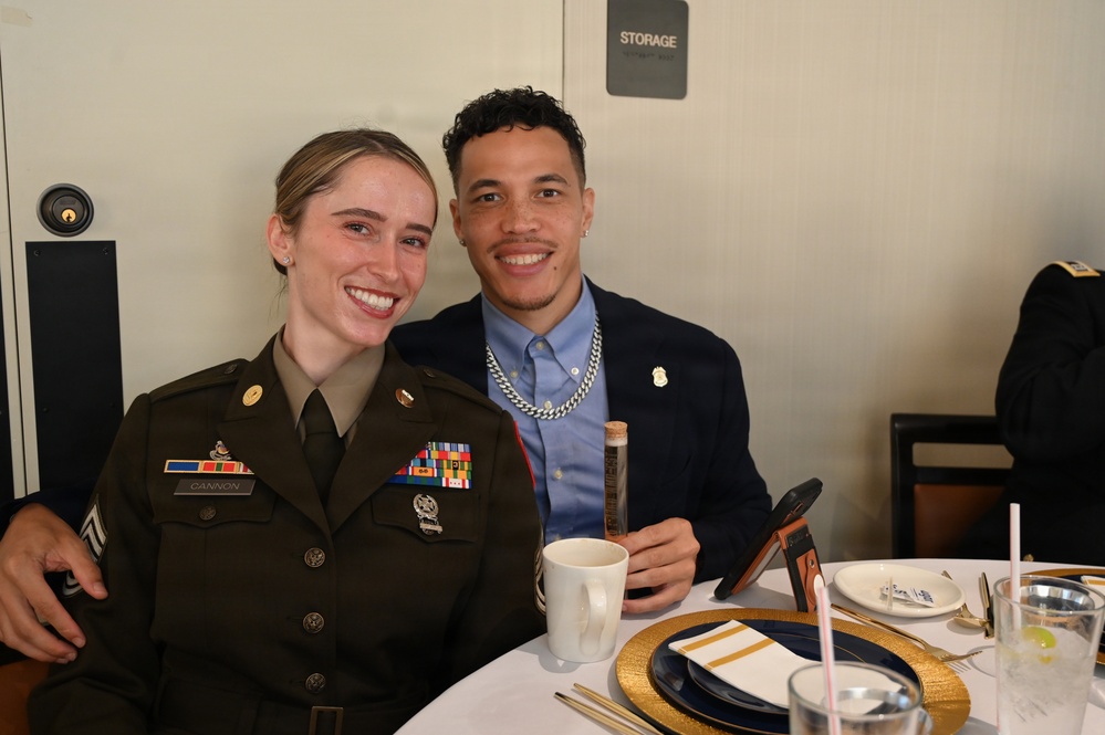 Two smiling individuals are seated at a formally set dining table, posing for a photo. The woman on the left is wearing a U.S. Army dress uniform with multiple service ribbons and a name tape reading 'CANNON,' holding the rank of Staff Sergeant. The man on the right is dressed in a navy blazer with a light blue shirt, silver chain necklace, and a badge pin.