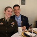 Two smiling individuals are seated at a formally set dining table, posing for a photo. The woman on the left is wearing a U.S. Army dress uniform with multiple service ribbons and a name tape reading 'CANNON,' holding the rank of Staff Sergeant. The man on the right is dressed in a navy blazer with a light blue shirt, silver chain necklace, and a badge pin.