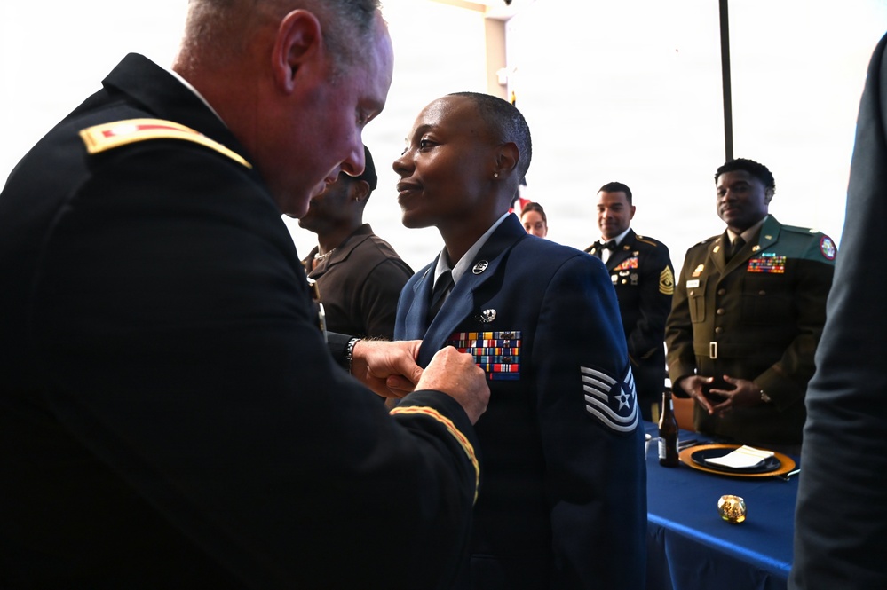 A military ceremony is taking place in which a U.S. Army officer is pinning a medal or insignia onto a U.S. Air Force Technical Sergeant in dress uniform. The recipient stands at attention while onlookers in various military uniforms observe the proceedings. A blue-draped table is visible in the background, consistent with a formal awards or promotion ceremony.