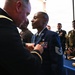 A military ceremony is taking place in which a U.S. Army officer is pinning a medal or insignia onto a U.S. Air Force Technical Sergeant in dress uniform. The recipient stands at attention while onlookers in various military uniforms observe the proceedings. A blue-draped table is visible in the background, consistent with a formal awards or promotion ceremony.