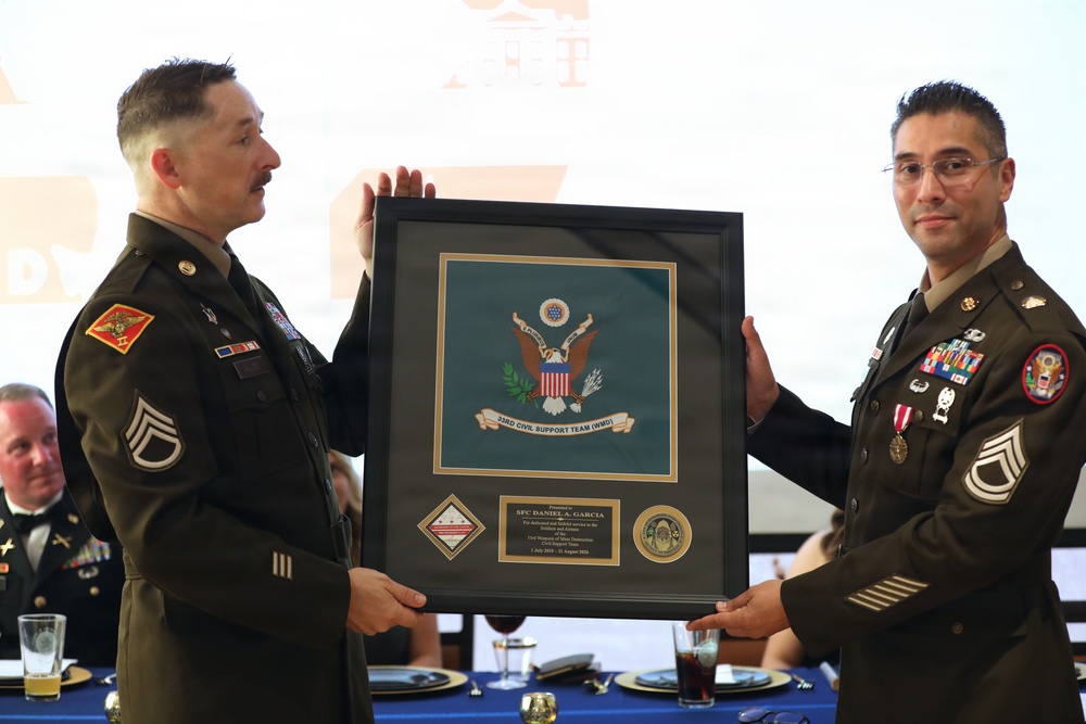 Two U.S. Army soldiers in dress uniforms are presenting a framed award or shadow box during what appears to be a formal military ceremony or banquet. The framed piece features the U.S. Army seal for the 33rd Civil Support Team (WMD) and is dedicated to SFC Daniel A. Garcia. The event is taking place in a banquet hall setting with other attendees visible in the background.