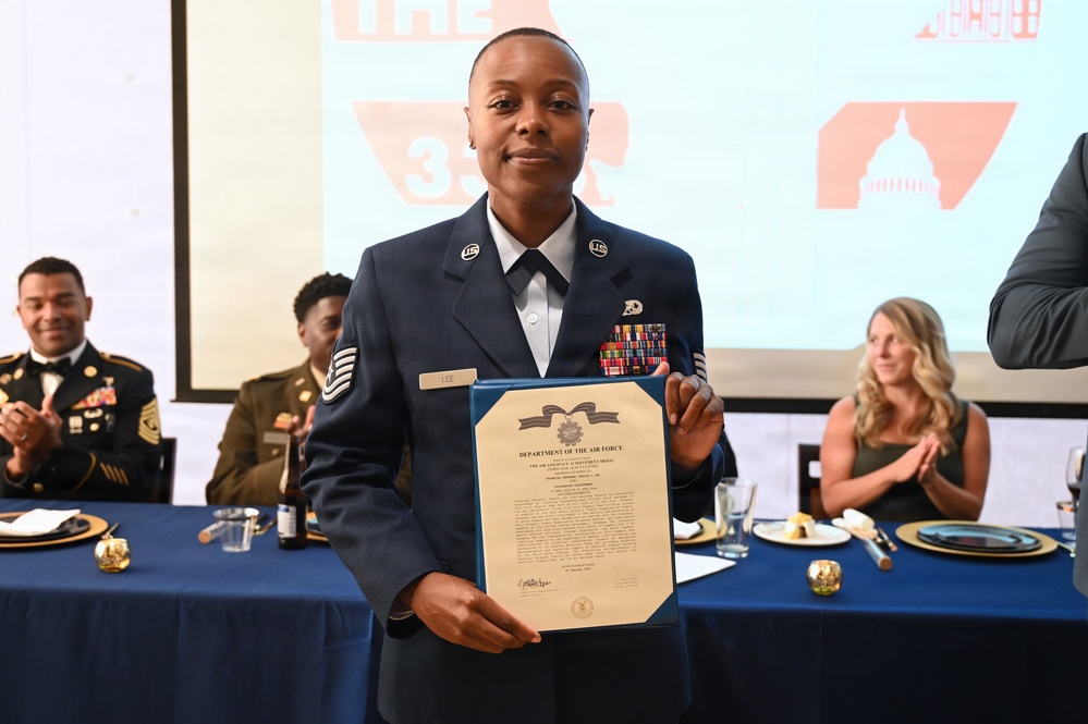 A U.S. Air Force service member named 'Lee' (visible on name tag) stands proudly holding a Department of the Air Force certificate, appearing to be the Air and Space Achievement Medal citation. The ceremony is taking place at a formal military banquet or awards dinner, with other military and civilian attendees seated at blue-draped tables in the background. Attendees are visibly applauding in recognition of the award recipient.