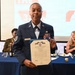 A U.S. Air Force service member named 'Lee' (visible on name tag) stands proudly holding a Department of the Air Force certificate, appearing to be the Air and Space Achievement Medal citation. The ceremony is taking place at a formal military banquet or awards dinner, with other military and civilian attendees seated at blue-draped tables in the background. Attendees are visibly applauding in recognition of the award recipient.