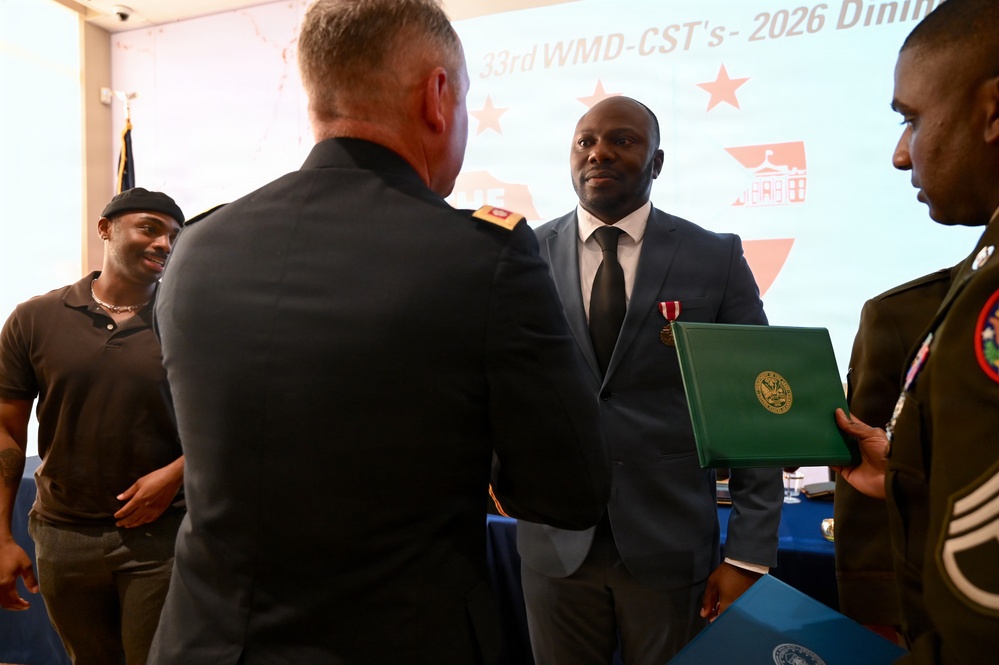 A formal military ceremony is taking place, likely a retirement or awards dinner associated with the '33rd WMD-CST's 2026 Dini...' event visible on the backdrop. A military officer in uniform is presenting or shaking hands with a civilian man in a dark suit who is wearing a red medal and holding a green official certificate folder. Additional attendees, including a uniformed soldier on the right, are present in the background.