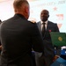 A formal military ceremony is taking place, likely a retirement or awards dinner associated with the '33rd WMD-CST's 2026 Dini...' event visible on the backdrop. A military officer in uniform is presenting or shaking hands with a civilian man in a dark suit who is wearing a red medal and holding a green official certificate folder. Additional attendees, including a uniformed soldier on the right, are present in the background.