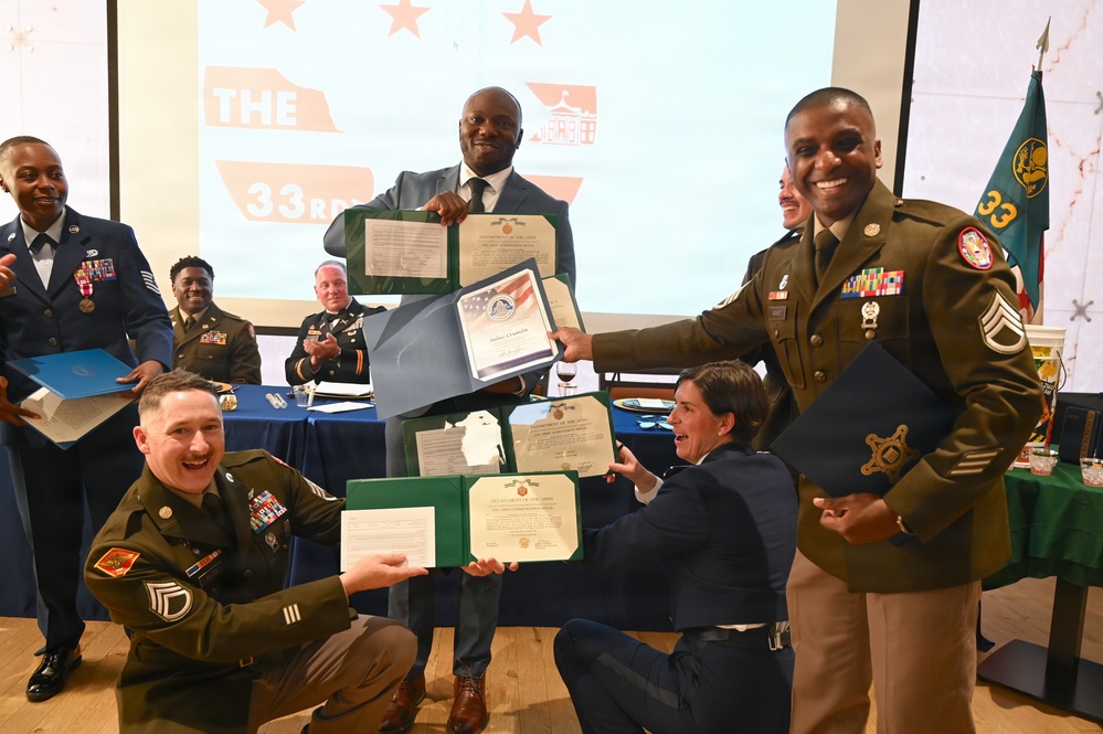 A group of military personnel in uniform are posing together at what appears to be an awards or recognition ceremony, holding official documents in green and blue folders. The event appears to be related to the '33rd' unit or occasion, as indicated by the presentation screen in the background. Multiple branches of the U.S. military are represented, with personnel displaying medals, ribbons, and rank insignia.