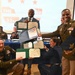 A group of military personnel in uniform are posing together at what appears to be an awards or recognition ceremony, holding official documents in green and blue folders. The event appears to be related to the '33rd' unit or occasion, as indicated by the presentation screen in the background. Multiple branches of the U.S. military are represented, with personnel displaying medals, ribbons, and rank insignia.
