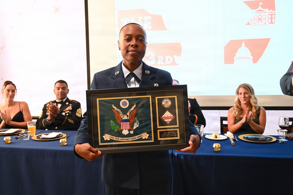 A military service member in dress uniform holds a framed commemorative plaque from the '33rd Civil Support Team (WMD)' at what appears to be a formal military banquet or awards ceremony. The plaque features the U.S. Great Seal and unit insignia. Attendees seated at blue-draped tables in the background are applauding the recipient.