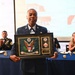 A military service member in dress uniform holds a framed commemorative plaque from the '33rd Civil Support Team (WMD)' at what appears to be a formal military banquet or awards ceremony. The plaque features the U.S. Great Seal and unit insignia. Attendees seated at blue-draped tables in the background are applauding the recipient.