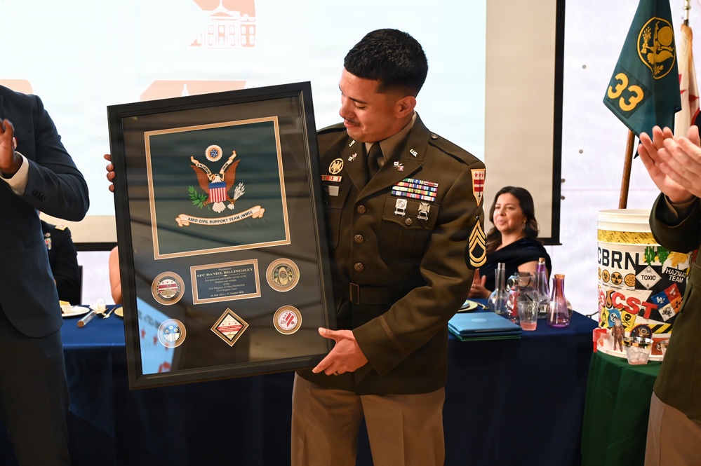 A U.S. Army soldier in dress uniform is holding a framed shadow box award at what appears to be a military ceremony or retirement event. The framed plaque bears the U.S. Army seal and reads '33rd Civil Support Team (WMD)' with the name 'SFC Daniel Billingsley' on the nameplate. Attendees are applauding in the background, and a unit guidon flag with the number '33' is visible to the right.
