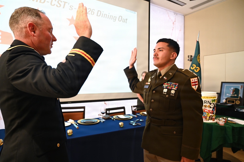 Two U.S. Army soldiers in dress uniforms are conducting a formal oath or re-enlistment ceremony, with both raising their right hands. The event appears to be taking place during a military dining-out ceremony, as indicated by the projected screen reading 'MD-CST's – 2026 Dining Out' and a formally set banquet table in the background. A flag with the number '33' is visible to the right side of the room.