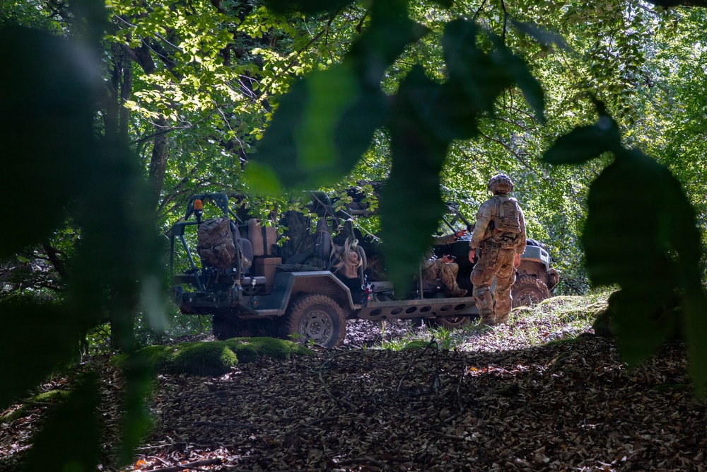 The image shows military personnel in a wooded area with a tactical light utility vehicle, photographed through foliage creating a natural frame. At least two soldiers in camouflage uniforms and helmets are visible near the heavily loaded vehicle. The scene appears to depict a field operation or training exercise in a dense forest environment.