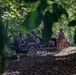 The image shows military personnel in a wooded area with a tactical light utility vehicle, photographed through foliage creating a natural frame. At least two soldiers in camouflage uniforms and helmets are visible near the heavily loaded vehicle. The scene appears to depict a field operation or training exercise in a dense forest environment.