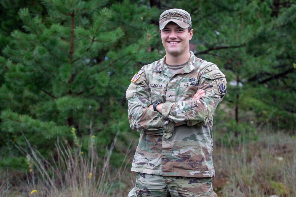 A U.S. Army soldier named Scaccia stands with arms crossed in an outdoor wooded setting, smiling at the camera. The soldier is wearing Operational Camouflage Pattern (OCP) uniform with visible name tape, U.S. Army tape, rank insignia, and unit patches. The image appears to be a formal or semi-formal military portrait taken in a natural environment.