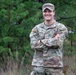 A U.S. Army soldier named Scaccia stands with arms crossed in an outdoor wooded setting, smiling at the camera. The soldier is wearing Operational Camouflage Pattern (OCP) uniform with visible name tape, U.S. Army tape, rank insignia, and unit patches. The image appears to be a formal or semi-formal military portrait taken in a natural environment.