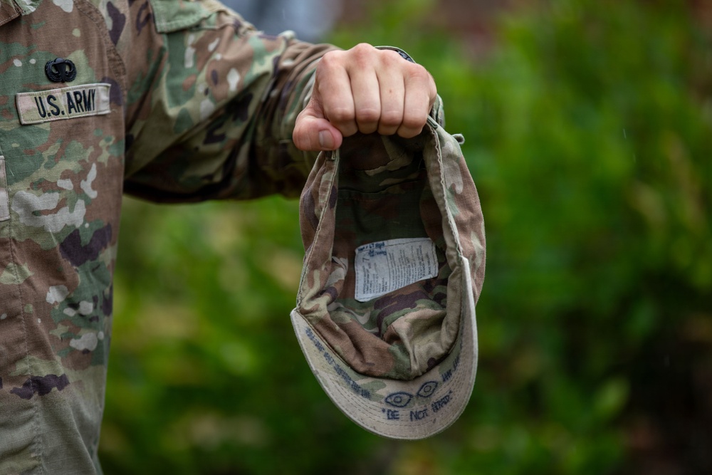 A U.S. Army soldier in Operational Camouflage Pattern (OCP) uniform holds an inverted patrol cap by its crown, revealing the interior lining and a white care/size label. The cap appears to have handwritten markings on the interior brim, including what appears to read 'DO NOT IRON' or similar text. The background shows blurred green foliage, suggesting an outdoor setting.