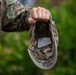A U.S. Army soldier in Operational Camouflage Pattern (OCP) uniform holds an inverted patrol cap by its crown, revealing the interior lining and a white care/size label. The cap appears to have handwritten markings on the interior brim, including what appears to read 'DO NOT IRON' or similar text. The background shows blurred green foliage, suggesting an outdoor setting.