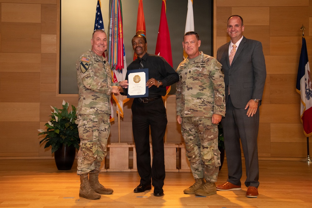 Four men pose for a formal award or recognition ceremony in what appears to be a military installation. Two individuals are wearing U.S. Army OCP (Operational Camouflage Pattern) uniforms, one man is in civilian business casual attire holding a framed certificate or award, and one man is in a business suit. Multiple military flags and unit colors are displayed in the background.