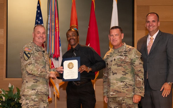 Four men pose for a formal award or recognition ceremony in what appears to be a military installation. Two individuals are wearing U.S. Army OCP (Operational Camouflage Pattern) uniforms, one man is in civilian business casual attire holding a framed certificate or award, and one man is in a business suit. Multiple military flags and unit colors are displayed in the background.