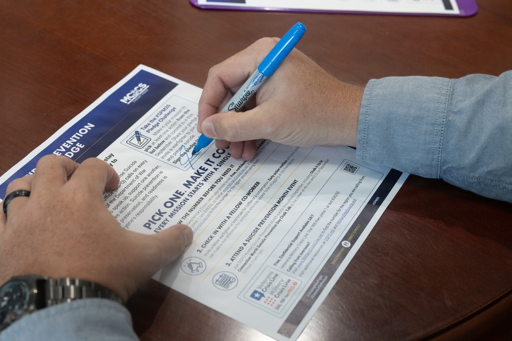 A person is signing a Suicide Prevention Pledge document on a wooden table using a blue Sharpie marker. The document, associated with MCCS (Marine Corps Community Services), encourages participants to take action during Suicide Prevention Month. The pledge includes options such as checking in with a fellow coworker and attending a suicide prevention month event.