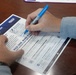 A person is signing a Suicide Prevention Pledge document on a wooden table using a blue Sharpie marker. The document, associated with MCCS (Marine Corps Community Services), encourages participants to take action during Suicide Prevention Month. The pledge includes options such as checking in with a fellow coworker and attending a suicide prevention month event.