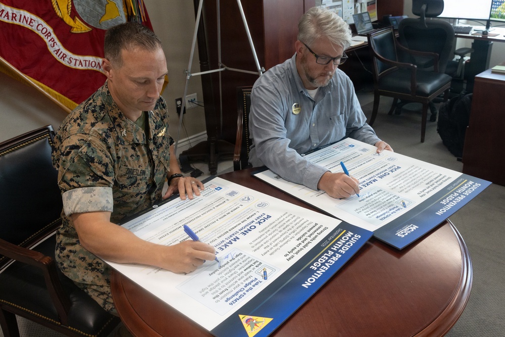 Two individuals are signing large pledge documents at a conference table in what appears to be a military office. One person is wearing U.S. Marine Corps camouflage uniform and the other is in civilian attire. The documents are identified as 'Suicide Prevention Month Pledge' forms associated with MCCS (Marine Corps Community Services).