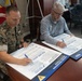 Two individuals are signing large pledge documents at a conference table in what appears to be a military office. One person is wearing U.S. Marine Corps camouflage uniform and the other is in civilian attire. The documents are identified as 'Suicide Prevention Month Pledge' forms associated with MCCS (Marine Corps Community Services).