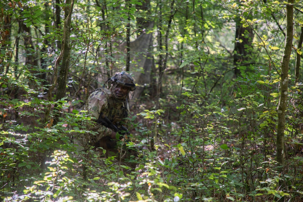 A soldier in full camouflage gear and helmet moves through dense woodland vegetation. The individual is partially obscured by surrounding foliage, suggesting a tactical movement or patrol exercise in a forested training environment. The soldier appears to be carrying a weapon and wearing a full combat kit.