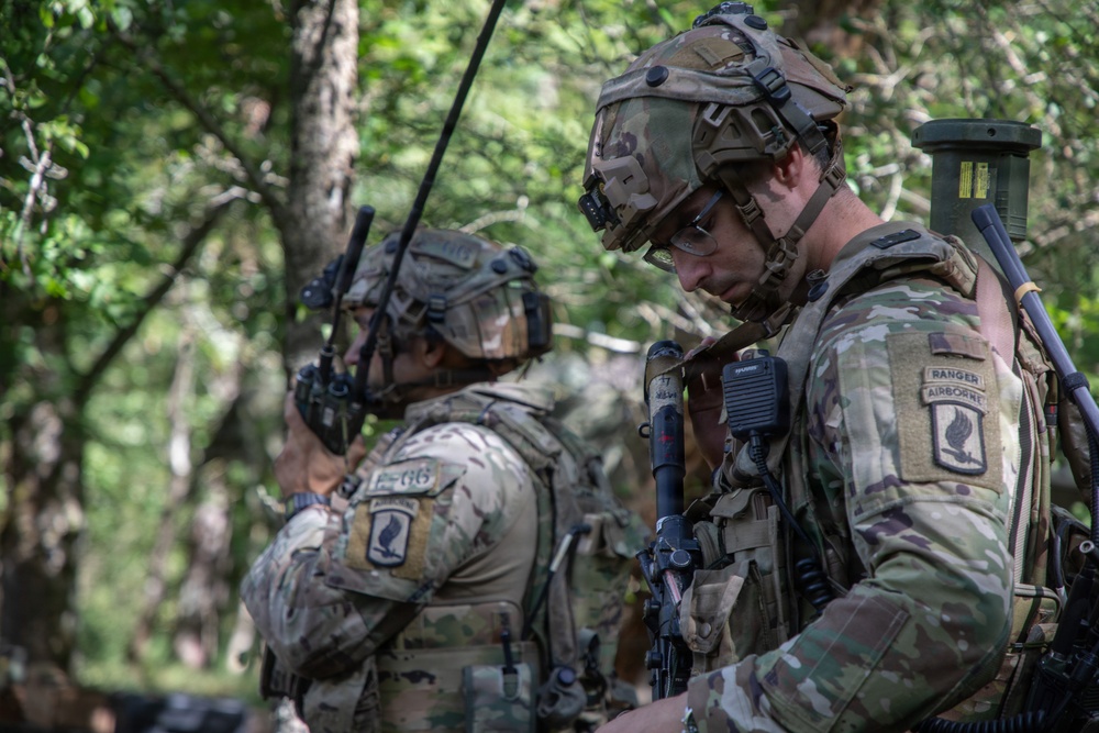 The image shows at least three U.S. Army soldiers in a wooded outdoor environment, wearing Operational Camouflage Pattern (OCP) uniforms and full tactical gear. The foreground soldier displays a 'Ranger' and 'Airborne' tab along with a 173rd Airborne Brigade patch, indicating elite airborne infantry unit affiliation. The soldiers appear to be conducting a field exercise or operation involving radio communications.
