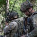 The image shows at least three U.S. Army soldiers in a wooded outdoor environment, wearing Operational Camouflage Pattern (OCP) uniforms and full tactical gear. The foreground soldier displays a 'Ranger' and 'Airborne' tab along with a 173rd Airborne Brigade patch, indicating elite airborne infantry unit affiliation. The soldiers appear to be conducting a field exercise or operation involving radio communications.