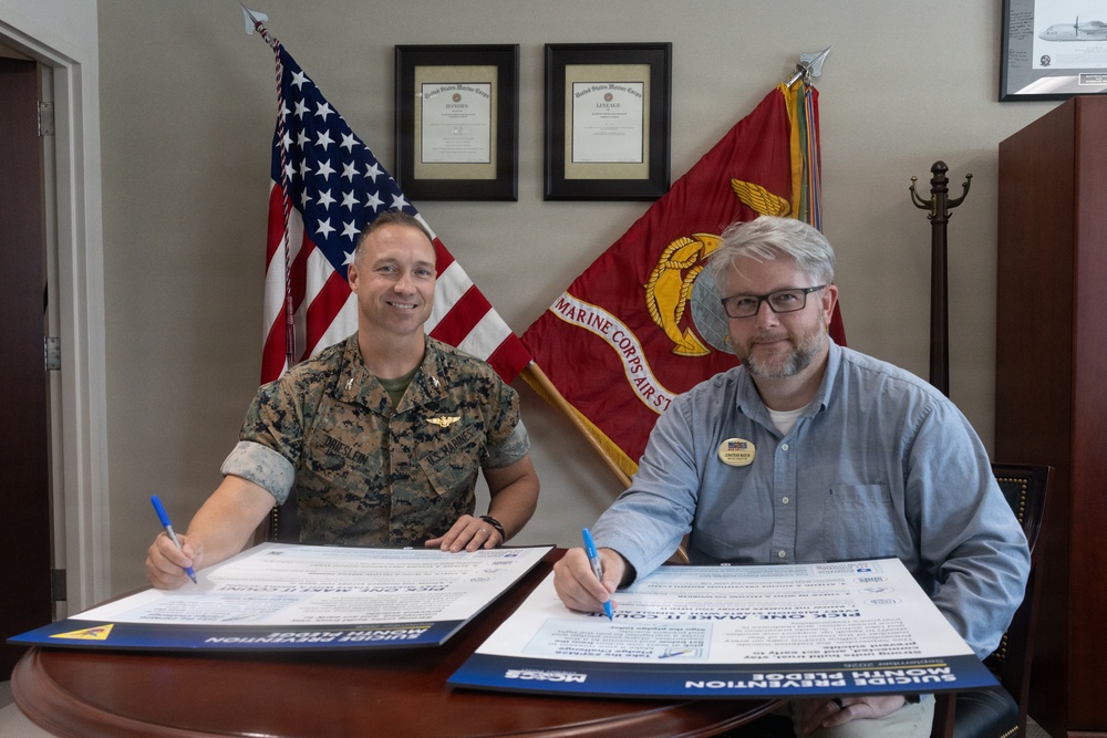 Two individuals are seated at a desk signing documents identified as 'Suicide Prevention Month Pledge' forms. One person is a U.S. Marine in camouflage uniform, and the other is a civilian wearing a blue button-down shirt with a name badge. The setting appears to be a military office with an American flag and Marine Corps Air Station flag visible in the background.