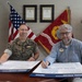 Two individuals are seated at a desk signing documents identified as 'Suicide Prevention Month Pledge' forms. One person is a U.S. Marine in camouflage uniform, and the other is a civilian wearing a blue button-down shirt with a name badge. The setting appears to be a military office with an American flag and Marine Corps Air Station flag visible in the background.