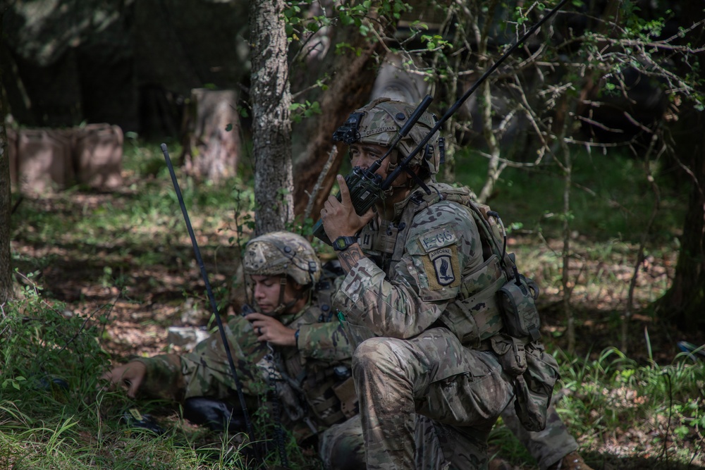 Two U.S. Army soldiers in MultiCam (OCP) camouflage uniforms are positioned in a wooded area conducting radio communications. The standing soldier is using a handheld radio while the seated soldier operates a separate radio system with a visible antenna. Unit patches indicate affiliation with the 173rd Airborne Brigade.
