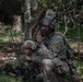 Two U.S. Army soldiers in MultiCam (OCP) camouflage uniforms are positioned in a wooded area conducting radio communications. The standing soldier is using a handheld radio while the seated soldier operates a separate radio system with a visible antenna. Unit patches indicate affiliation with the 173rd Airborne Brigade.