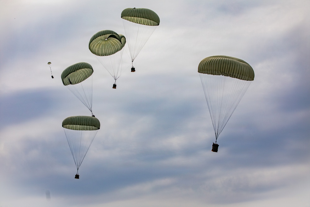 The image shows multiple military cargo parachutes descending through an overcast sky. All parachutes are olive/sage green in color and appear to be carrying suspended loads or equipment packages beneath them. This appears to be a military airdrop or supply delivery operation.
