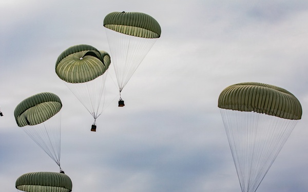 The image shows multiple military cargo parachutes descending through an overcast sky. All parachutes are olive/sage green in color and appear to be carrying suspended loads or equipment packages beneath them. This appears to be a military airdrop or supply delivery operation.
