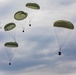 The image shows multiple military cargo parachutes descending through an overcast sky. All parachutes are olive/sage green in color and appear to be carrying suspended loads or equipment packages beneath them. This appears to be a military airdrop or supply delivery operation.