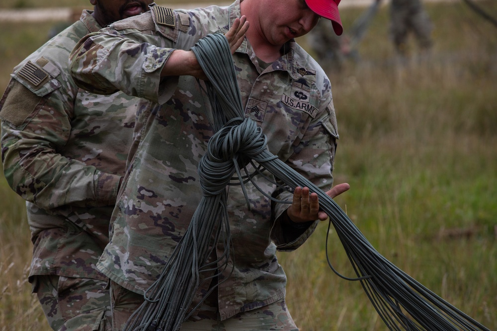 Two U.S. Army soldiers in OCP (Operational Camouflage Pattern) uniforms are handling a large bundle of parachute suspension lines or similar cordage in an outdoor field setting. The soldier in the foreground, wearing a red cap and displaying a 'U.S. ARMY' tape and sergeant rank insignia, is carrying the bundled lines while the other soldier appears to be assisting or directing. The activity appears to be post-parachute jump recovery or rigging operations.