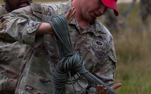 Two U.S. Army soldiers in OCP (Operational Camouflage Pattern) uniforms are handling a large bundle of parachute suspension lines or similar cordage in an outdoor field setting. The soldier in the foreground, wearing a red cap and displaying a 'U.S. ARMY' tape and sergeant rank insignia, is carrying the bundled lines while the other soldier appears to be assisting or directing. The activity appears to be post-parachute jump recovery or rigging operations.