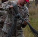 Two U.S. Army soldiers in OCP (Operational Camouflage Pattern) uniforms are handling a large bundle of parachute suspension lines or similar cordage in an outdoor field setting. The soldier in the foreground, wearing a red cap and displaying a 'U.S. ARMY' tape and sergeant rank insignia, is carrying the bundled lines while the other soldier appears to be assisting or directing. The activity appears to be post-parachute jump recovery or rigging operations.