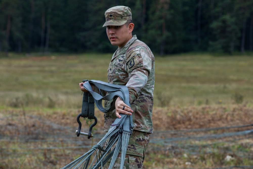 A U.S. Army soldier in Operational Camouflage Pattern (OCP) uniform is handling rigging equipment, including straps and suspension lines with a metal shackle, in an open field. The soldier appears to be managing or recovering parachute or sling load rigging components. The setting appears to be a military training area bordered by a tree line.