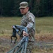 A U.S. Army soldier in Operational Camouflage Pattern (OCP) uniform is handling rigging equipment, including straps and suspension lines with a metal shackle, in an open field. The soldier appears to be managing or recovering parachute or sling load rigging components. The setting appears to be a military training area bordered by a tree line.