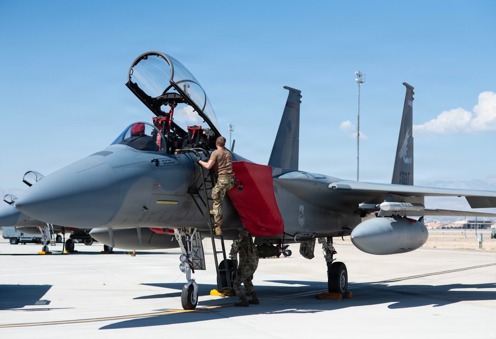 A military aircraft technician in camouflage uniform is performing maintenance or pre-flight checks on an F-15 Eagle fighter jet on a flight line. The canopy is open and a maintenance ladder with a red cover is positioned at the cockpit. The scene appears to be at a U.S. Air Force base under clear blue skies.