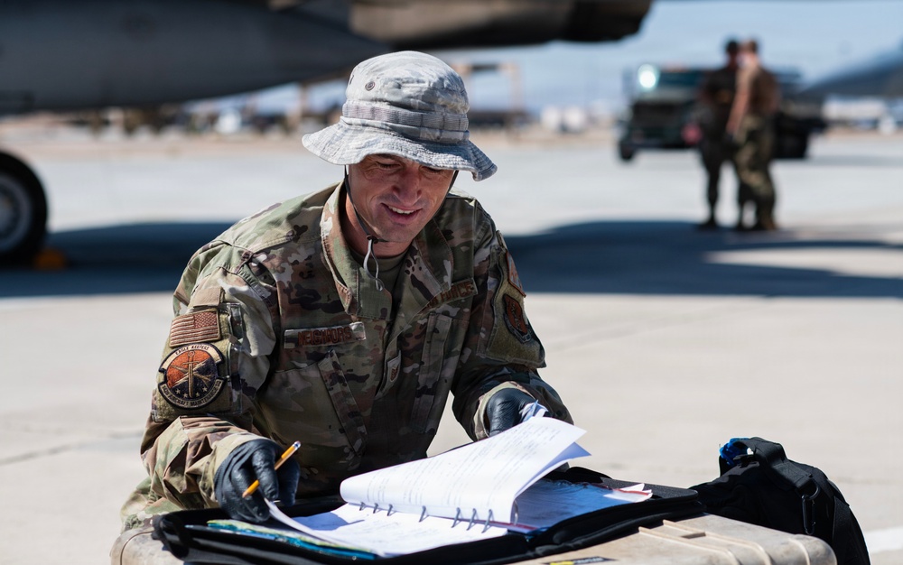 A U.S. Air Force service member in OCP (Operational Camouflage Pattern) uniform is reviewing documents on a flight line, likely conducting pre-flight or maintenance checks. He is wearing a boonie hat and black gloves while holding a pencil over a binder or clipboard. The setting appears to be a desert air base with aircraft and other personnel visible in the background.