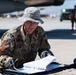 A U.S. Air Force service member in OCP (Operational Camouflage Pattern) uniform is reviewing documents on a flight line, likely conducting pre-flight or maintenance checks. He is wearing a boonie hat and black gloves while holding a pencil over a binder or clipboard. The setting appears to be a desert air base with aircraft and other personnel visible in the background.