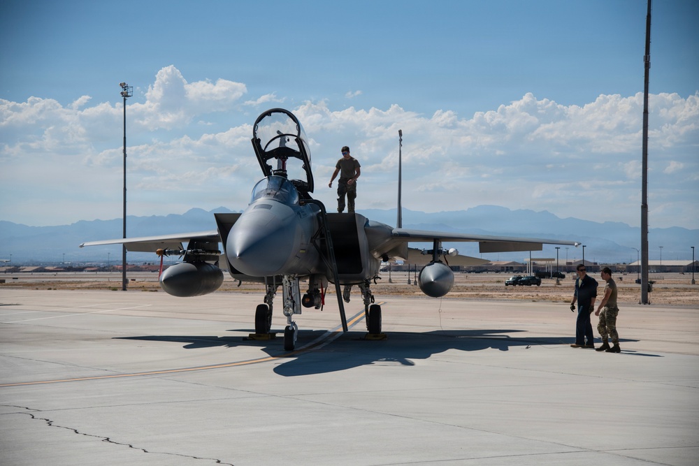 A military fighter jet, consistent with an F-15 Eagle, is parked on a concrete flight line at what appears to be a U.S. Air Force base. Ground crew personnel are conducting post-flight or pre-flight maintenance checks, with one individual standing on top of the aircraft near the open cockpit canopy. The background reveals a desert airbase environment with mountains visible on the horizon.
