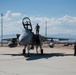 A military fighter jet, consistent with an F-15 Eagle, is parked on a concrete flight line at what appears to be a U.S. Air Force base. Ground crew personnel are conducting post-flight or pre-flight maintenance checks, with one individual standing on top of the aircraft near the open cockpit canopy. The background reveals a desert airbase environment with mountains visible on the horizon.